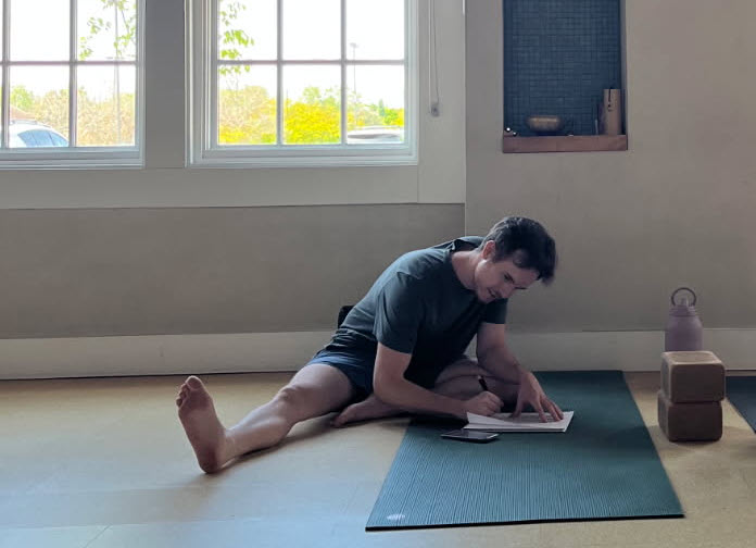 Tyler sitting on a green yoga mat in a sunlit studio, writing in a notebook with a phone beside him.